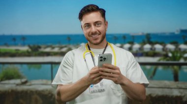 Deniz kenarındaki gezinti güvertesinde akıllı telefon kullanırken gülümseyen sakallı genç bir doktor, steteskop tutuyor, çarpıcı mavi deniz zeminine karşı modern sağlık hizmetleri iletişimi sergiliyor..