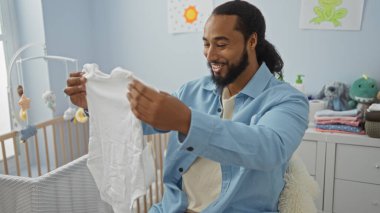 Bebek odasında bebek kıyafetleri tutan bir adam gülümserken oyuncak dolu bir beşikte yumuşak pastel renkte bir evde oturuyor..