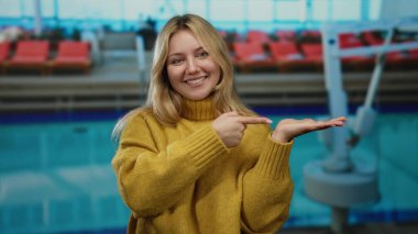 Havuz başında gülümseyen bir kadın açık eli işaret ediyor sarı süveter giyiyor açık havada rahat bir atmosfer..