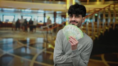 Bir otel lobisinde Rus rublesi banknotları tutan adam gülümseyen bir ifade ile, kapalı arka planda bulanık, zenginlik ve modern yaşam tarzı vurguluyor.