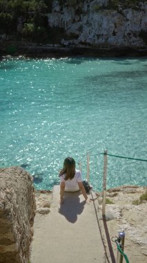 Güneşli bir günde Mallorca 'da turkuaz suları olan Cala Lombars plajının keyfini çıkaran kadın, Akdeniz sahilinin sükunetini ve doğal güzelliğini vurguluyor..