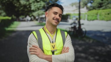 Yansıtıcı yelek giyen genç bir adam parkta gönüllü olarak çalışıyor, kendine güvenen bir ifadeyle kollarını kavuşturuyor, güneşli bir günde açık havada duruyor, bulanık yeşil bir arka planı var..