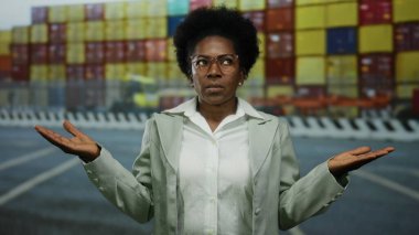 Kargo konteynırlarının önünde açık havada duran, kafası karışmış bir ifade ve kolları açık bir kadın, hareketli sahil ortamında belirsiz bir an sergiliyor..