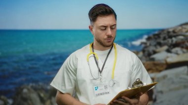 Deniz kenarındaki panoya bakan sakallı genç doktor. Arka planda okyanus var. Açık havada steteskop takıyor.