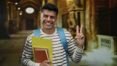 Müzede elinde defterlerle gülümseyen genç bir adam çizgili bir tişört ve mavi bir sırt çantası giymiş, tarihi bir sanat eserinde barış işareti taşıyor.