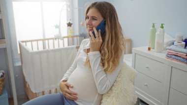Hamile kadın rahat yatak odasında oturmuş telefonla konuşuyor, gülümsüyor, arka planda bebek beşiği, yumuşak ışık ve ev dekoruyla çevrili, sakin, beklenen bir atmosfer yaratıyor..
