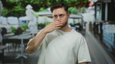 Şehir terasındaki sakallı genç adam, şehir restoranındaki masaların ve yeşil ağaçların yer aldığı kalabalık caddedeki kafe yakınlarında sürpriz bir şekilde tepki veriyor..