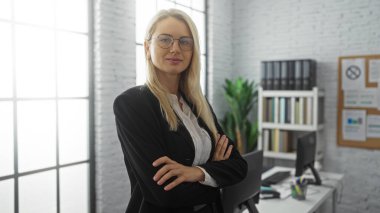 Kadın, modern ofiste kollarını kavuşturarak duruyor. Etrafı kitaplık ve masalarla çevrili profesyonellik ve liderlik yayıyor. Çalışma ortamını büyütüyor..