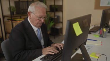 Modern ofis içinde bir bilgisayarda çalışan takım elbiseli kıdemli bir adam, belgeleri okumaya ve görevleri düzenlemeye odaklanmış..