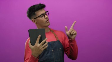 Gözlüklü genç adam elinde tablet, parlak pembe arka plan önlüğü ile derin düşünce ve dikkat çekmeyen bir şeyi işaret ediyor..