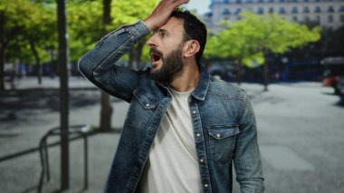 Bir şehir caddesinde duran İspanyol sakallı adam, şaşırmış ve düşünceli görünüyor, bir kot ceket ve canlı yeşil ağaçların altında beyaz gömlek giyiyor şehir arka planında.