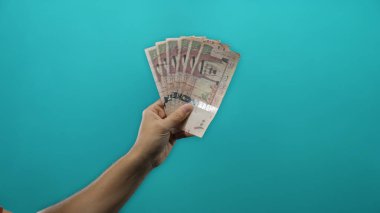 Suudi riyal banknotlarını canlı, mavi bir zemin üzerinde tutan bir adamın el ele tutuşması, izole edilmiş bir ortamda para birimi ve finans temalarını vurguluyor.