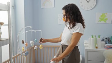 Yatak odasında duran bir kadın, rahat bir ev ortamında bebek beşiği üzerine cep telefonunu ayarlıyor. Bakımlı ve sade bir atmosfer öneriyor..