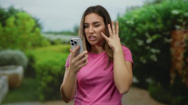 Parktaki akıllı telefonuna sinirlenmiş bir kadın açık havada, açık havada, yemyeşil bir ortamda, pembe bir tişört giyip, hoşnutsuzluğunu ifade ediyor..