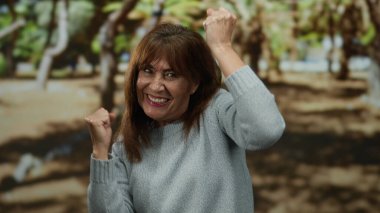 Üst düzey İspanyol kadın açık hava parkında kollarını kaldırarak mutlu bir şekilde kutlama yapıyor. Etrafı bulanık yapraklarla çevrili ve parlak bir günde rahat bir kazak giyiyor..