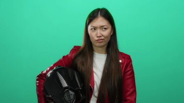 Kırmızı ceketli kadın yeşil arka planda miğfer tutuyor, güçlü bir ifade ve uzun saçlar sergiliyor, kendinden emin, şık bir hava yayıyor, modern, kentsel, kadın varlığını sembolize ediyor.