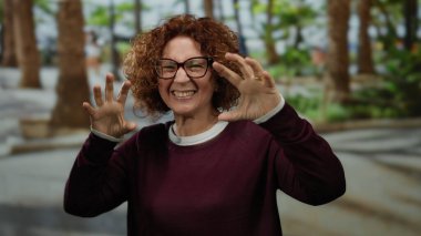 Kadın, arka planda ağaçlarla güneşli bir parkta neşe içinde poz veriyor, gözlük takıyor, bordo bir kazak giyiyor, dışarıda neşe ve mutluluğu ifade ediyor..