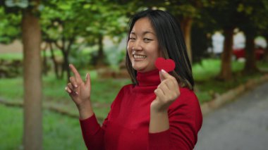 Parkta elinde kağıt kalple gülümseyen, kırmızı süveter giyen, doğa ve ağaçlarla çevrili, huzurlu bir Çin ortamında neşe ve pozitifliğini ifade eden bir kadın..