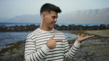 Sahilde duran genç adam başparmaklarını kaldırarak ve açık bir el göstererek arka planda dağlar ve bulutlu gökyüzü altında dalgalar olan çizgili tişört giyerek kıyıda duruyor.