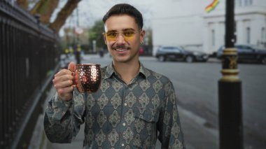 Şablon gömlekli ve sarı gözlüklü genç İspanyol bir adam şehir sokaklarında bir bakır bardak tutuyor, açık şehir ortamında neşeli bir hava yayıyor..