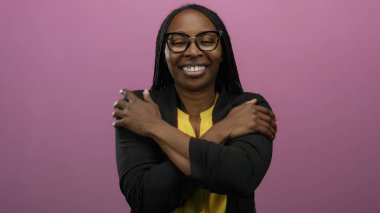 Kollarını sıcak bir şekilde kavuşturarak gülümseyen bir kadın, soyutlanmış pembe arka plana karşı duran, sükuneti somutlaştıran ve kendine güvenen gözlüklü ve kendinden emin bir ifade takınan bir kadın..