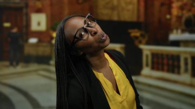 Gözlüklü Afrikalı Amerikalı bir kadın arka planda zarif bir dekor olan bir kilise ortamında neşeyle gülümsüyor..