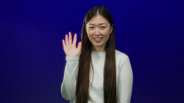 Mavi arka planda el sallayan ve gülümseyen bir kadın, dostça bir jest ve sıradan bir moda sergiliyor..