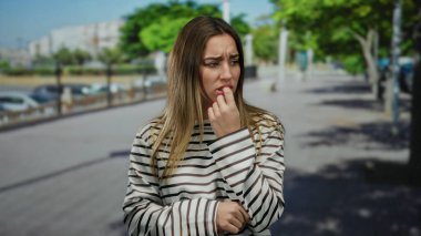 Çizgili tişört giyen kadın, arka planda yeşil ağaçlar ve binalar olan güneşli şehir bölgesinde sokakta endişeli görünüyor..