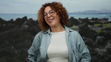 Açık havada bulutlu bir günde deniz kenarında esmer bir kadın neşeli bir şekilde gülümsüyor..