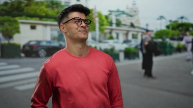 Kırmızı kazaklı genç adam şehir caddesinde özgüvenle gülümsüyor yemyeşil ve modern mimarisi arka planda bulanık..