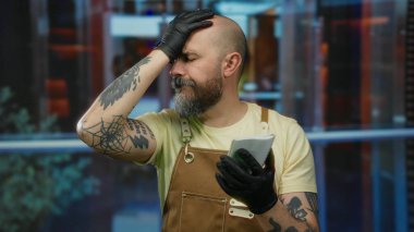 Otelde sakallı, not defteri tutan, eldivenli, önlüklü, evde dövmeli bir adam hayal kırıklığı ifadesi sergiliyor..