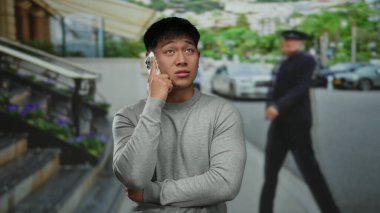 Gri kazaklı genç adam şehir caddesinde akıllı telefonla konuşuyor. Etrafı arabalar ve yeşilliklerle çevrili..