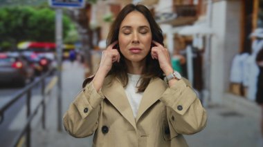 Bej ceketli bir kadın, gürültüyü engellemek için yoğun şehir caddesiyle çevrili kulaklarını kapatıyor. Bu da kentsel dikkat dağınıklığından kaynaklanan stres ve rahatsızlığı gösteriyor..