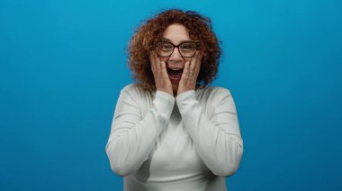 Gözlük takan bir kadın canlı mavi arka plan duvarına karşı şaşkınlık ve heyecan ifade ediyor. Görüntüde canlı ve çekici bir atmosfer yaratıyor..