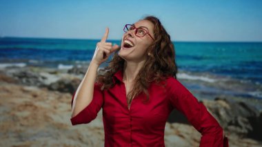 Kırmızı gömlekli kadın deniz kenarında neşeyle gülüyor, açık mavi deniz arka planına işaret ediyor, kumsaldaki mutluluk ve heyecanı somutlaştırıyor..