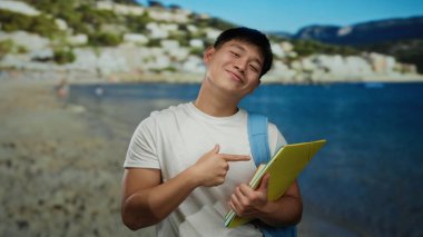 Deniz kenarında gülümseyen ve baş parmağını kaldıran genç bir adam bir klasör tutarken, bir sırt çantası takarken, arkasında açık mavi bir deniz olan kumsal manzarasının tadını çıkarıyor..
