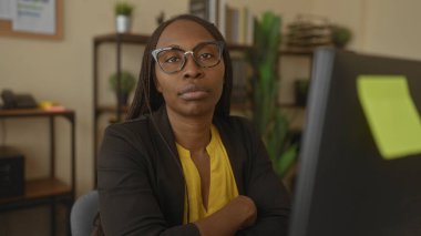 Kıvırcık saçlı, gözlüklü bir kadın ofis ortamında oturmuş, evde çalışırken düşünceli bir yüz ifadesi sergiliyor..