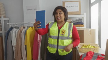 Kıyafet ve kutularla dolu bir bağış merkezinden yansıtıcı yelek giymiş bir kadın arıyor. Canlı bir toplum gönüllüsünü kapalı alanda sergiliyor..