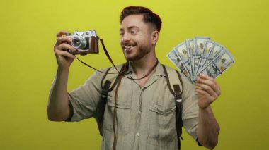 Sırt çantası ve kamera ile gülümseyen genç adam parlak sarı arka plana karşı bize dolar tutuyor başarılı bir seyahat fotoğrafçılığı deneyimi öneriyor