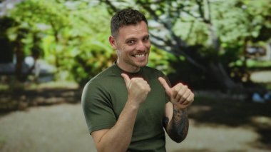 Parkta baş parmağıyla gülümseyen genç bir adam açık havada neşeli ve kendinden emin bir yüz ifadesi sergiliyor yemyeşil ağaçlarla çevrili aydınlık bir günde..