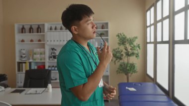 Yeşil üniformalı genç adam hastane kliniğinde elinde gözlüklerle dikiliyor. Profesyonel bir tıbbi ortamda pencereden dışarı bakıyor..