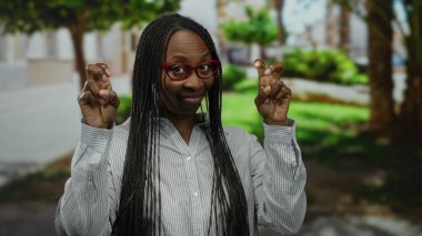 Kıvırcık saçlı bir kadın, şehir sokaklarında, gözlük ve çizgili bir gömlek takıp, açık hava ortamında neşeli ve kendinden emin bir ifade sergilerken parmak tırnak işareti yapıyor..
