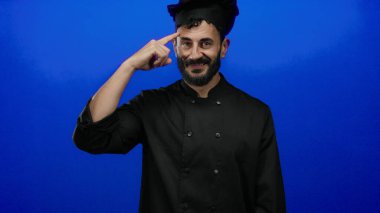 Siyah şef üniformalı İspanyol bir adam sakallı, parlak mavi bir arka plana karşı düşünceli, profesyonel bir yemek pişirme havası yayıyor..