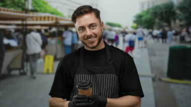 Önlüklü genç adam açık hava pazarında elinde hazır kahveyle şehir arka planında hareketli gülümsüyor ve sokak ortamında içki servisi yapan bir garson gibi gülümsüyor..