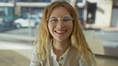 Ofis ortamında gülümseyen genç bir kadın modern bir iç mekan çalışma ortamında güven içinde poz verirken sarı saçlarını ve gözlüklerini öne çıkarıyor..