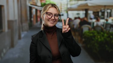 Şehir terasında gülümseyen bir kadın yeşil bitkilerle dolu bir restoranın ortasında gözlük ve spor ceket giyerek barış jesti yapıyor..