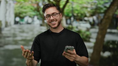 Sakallı ve gözlüklü genç adam, şehir caddesinde akıllı telefona bakarken gülümsüyor, arka planda ağaçlarla şehir merkezinin tadını çıkarıyor..