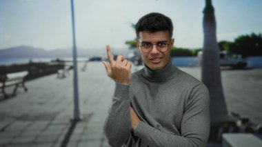 Gözlük takan genç adam, güneşli bir sahil kenarında, arka planda okyanus ve palmiye ağaçlarıyla düşünceli bir şekilde el kol hareketi yapıyor..