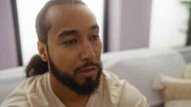 Oturma odasında sakallı, düşünceli ve rahat, modern dekorlu, kültürel geçmişi ve modern yaşam tarzını sergileyen genç bir adam.