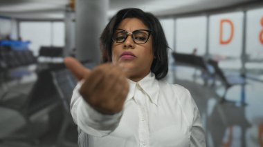 Havalimanı terminaline arka planda oturma ve işaret levhası olan, gözlüklü ve beyaz gömlekli bir kadın gelir, kapalı mekanda kendine güvenen bir davet ifadesi yakalar..